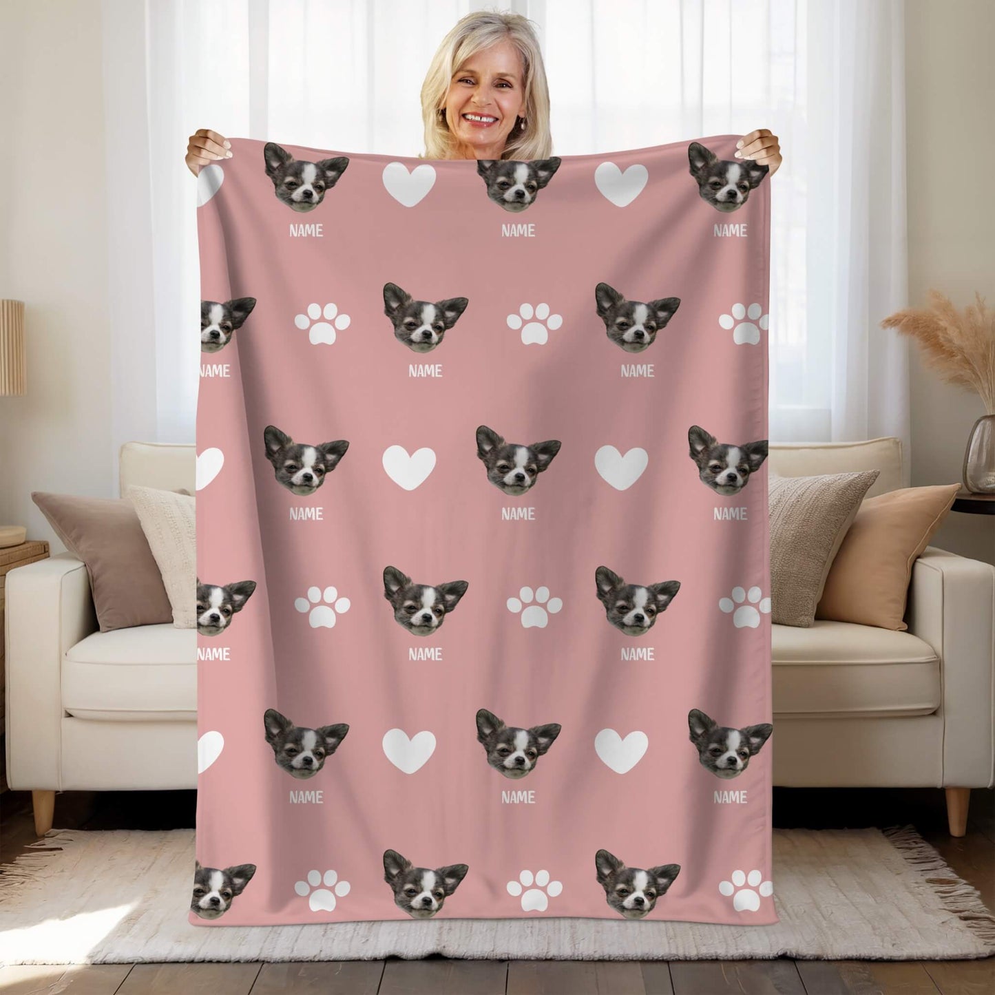 Person holding a pink blanket with dog faces and paw prints in a living room.