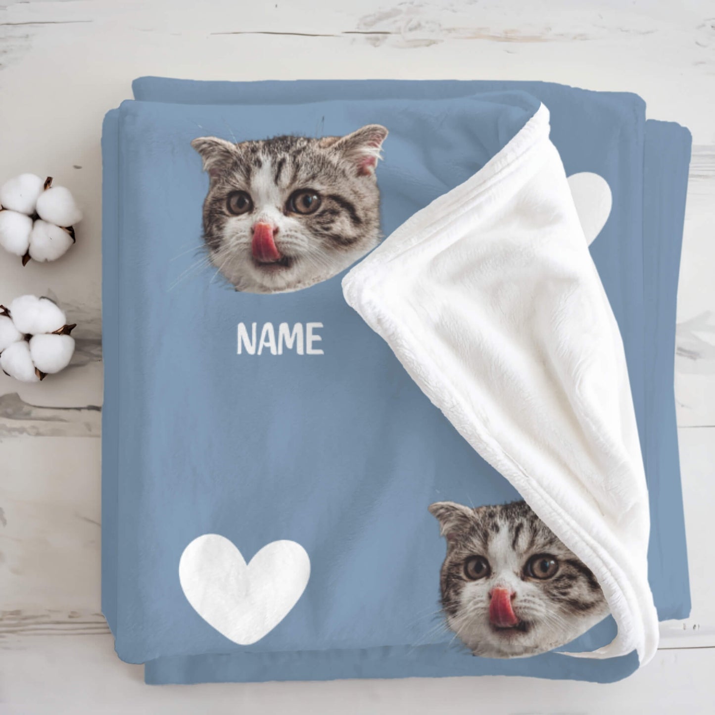 Folded blanket with cat face design and 'NAME' text on a wooden surface.